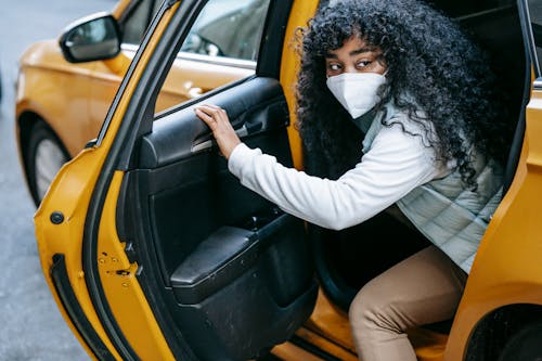 African American female with black hair in protective mask casual clothes getting out of yellow car and looking away in daytime