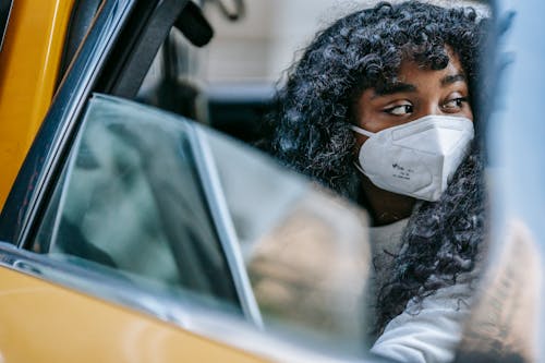 Black young lady in casual clothes and protective mask sitting in yellow cab with open door and looking away thoughtfully in daytime in city street