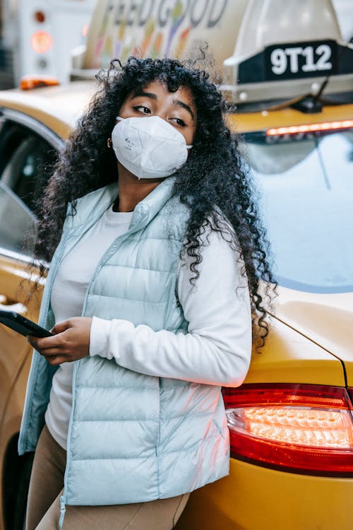 Young black lady wearing protective mask and casual clothes while leaning on yellow cab and browsing on phone in city street in daytime and looking away thoughtfully