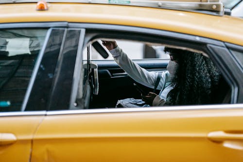 Young black lady wearing protective mask and casual clothes sitting with backpack in yellow cab while surfing on smartphone with open window in street in daytime in town