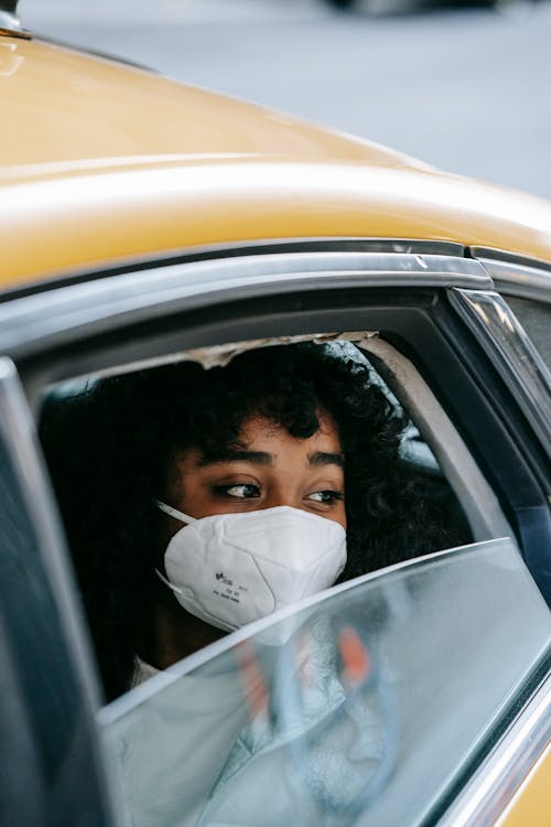 African American young lady in casual outfit and protective mask sitting in yellow taxi with open window and looking away thoughtfully in city street in daylight