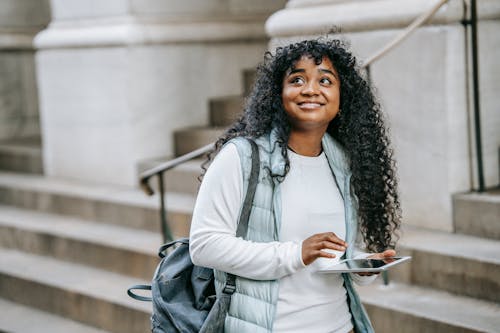 Happy young African American woman wearing casual clothes with backpack while using tablet on city street near building with stone stairs in daylight