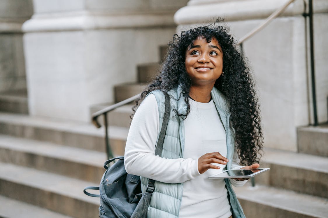 Free Happy young African American woman wearing casual clothes with backpack while using tablet on city street near building with stone stairs in daylight Stock Photo