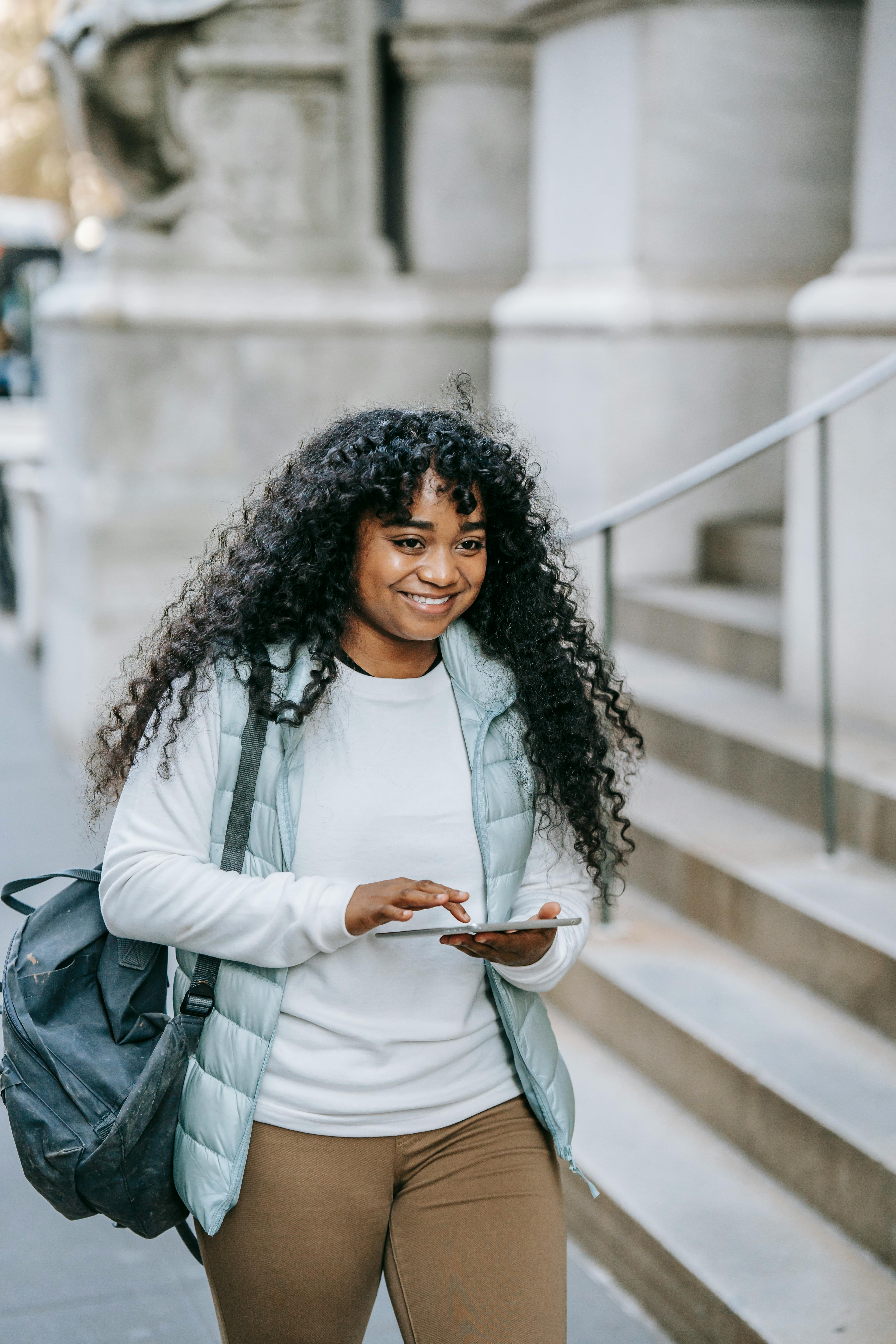 Smiling young black lady in casual outfit with backpack on city street with tablet near building with stone steps in daytime