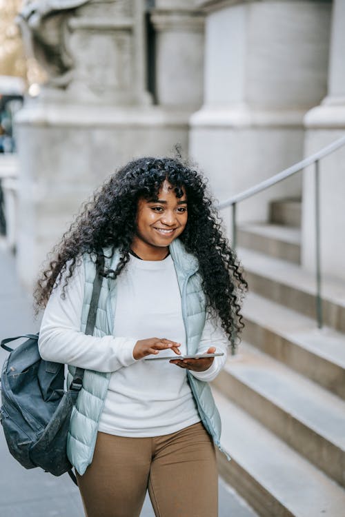 Free Smiling young black lady in casual outfit with backpack on city street with tablet near building with stone steps in daytime Stock Photo