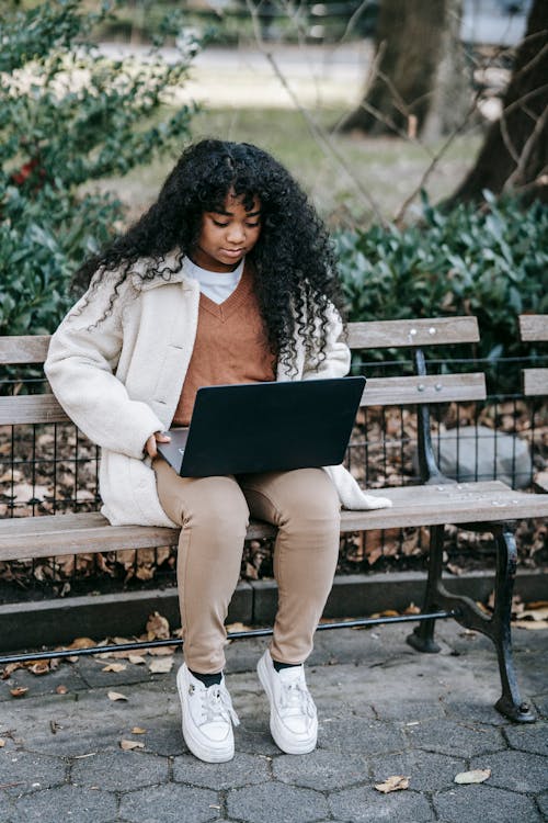 Full body of thoughtful young African American lady in casual outfit sitting on wooden bench in park while surfing on netbook near plants and trees near fallen leaves in daylight