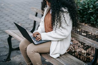 Crop concentrated African American female in warm jacket working on modern netbook and sitting on wooden bench in spring city park