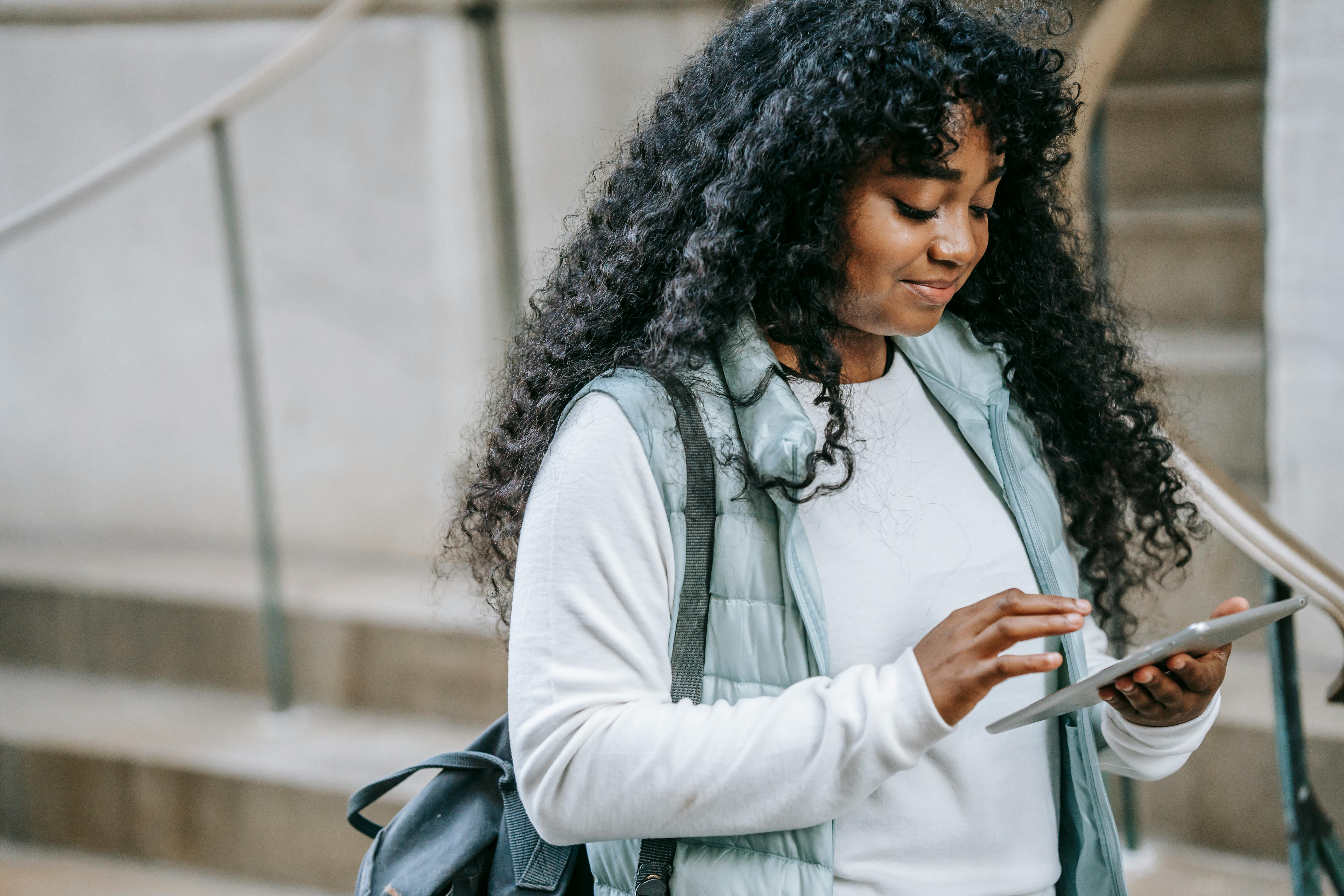 Content African American female with long curly hair wearing warm jacket browsing modern tablet while standing on city street
