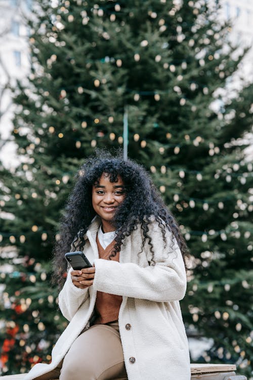 Smiling black woman using smartphone near decorated Christmas tree