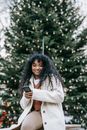 Smiling black woman using smartphone near decorated Christmas tree