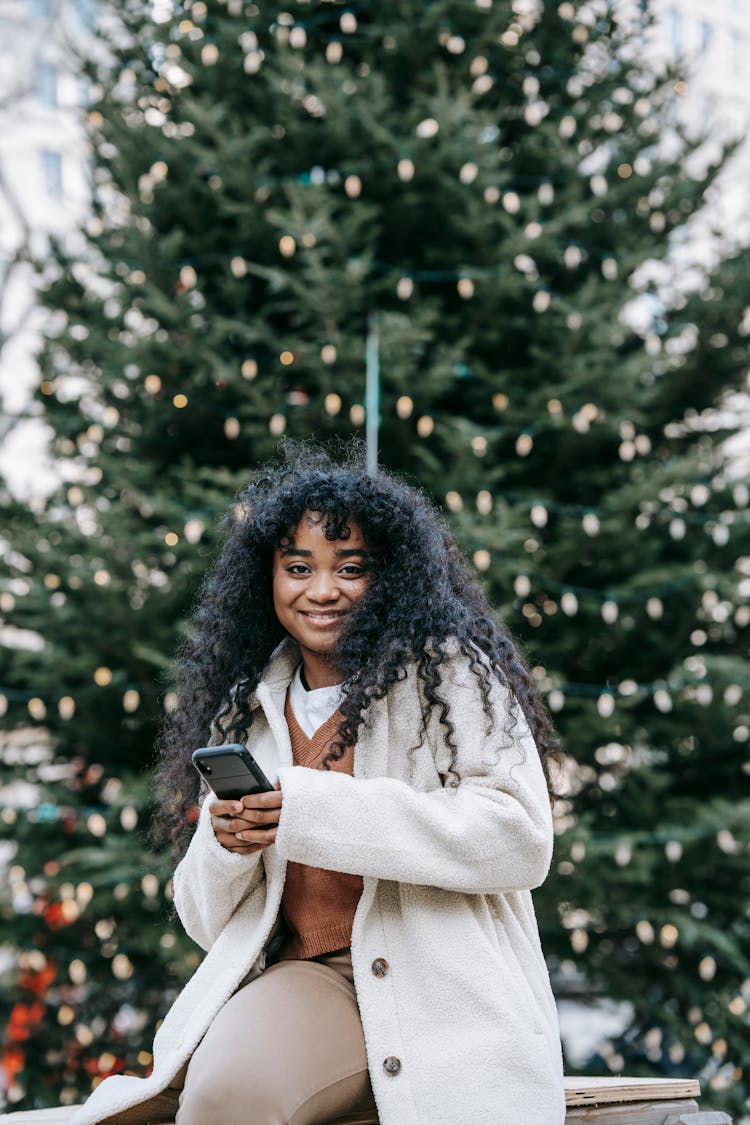 Smiling Black Woman Using Smartphone Near Decorated Christmas Tree