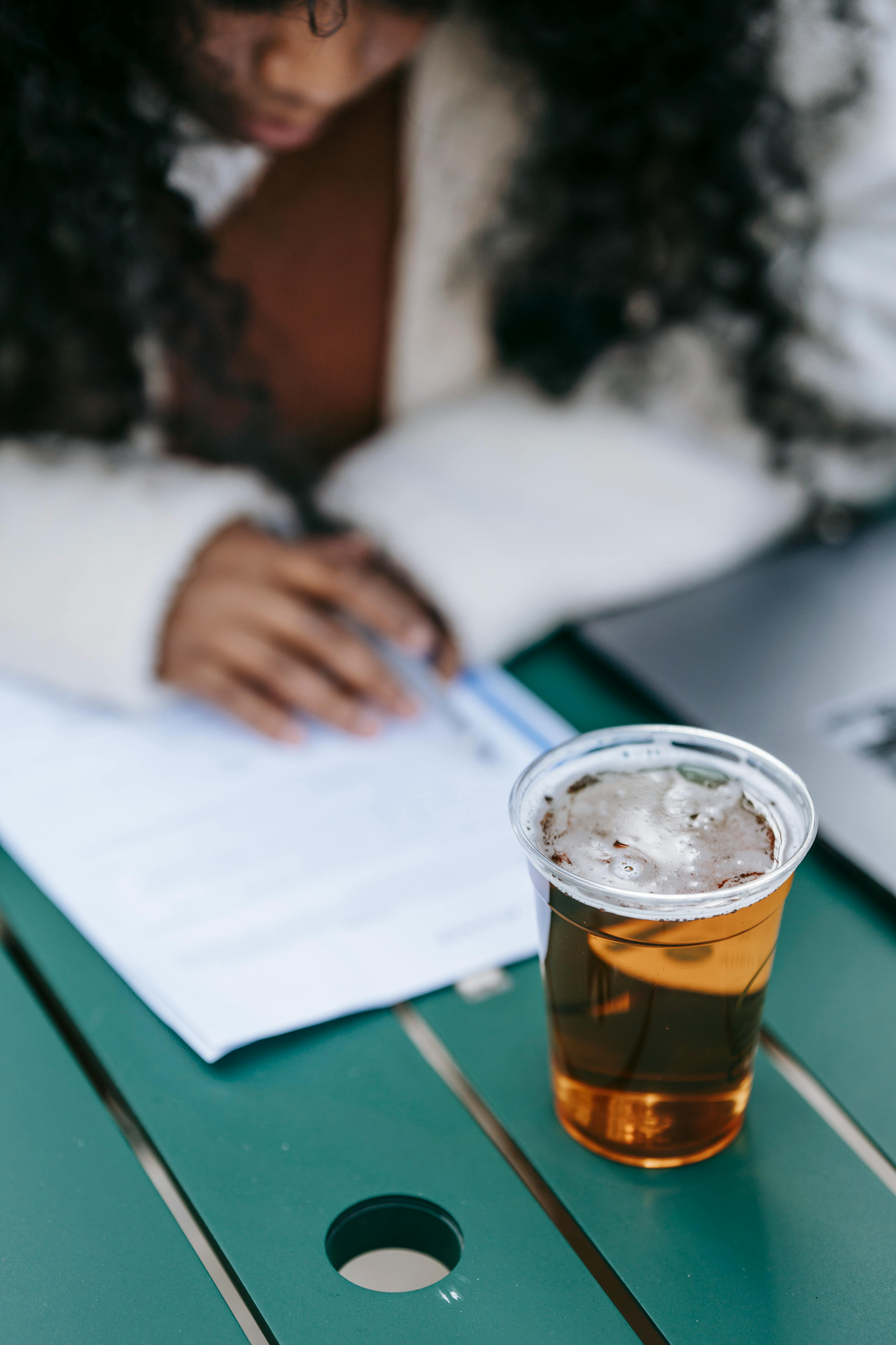 Crop concentrated African American female with long curly hair doing paperwork while sitting at table with glass of cold beer and laptop