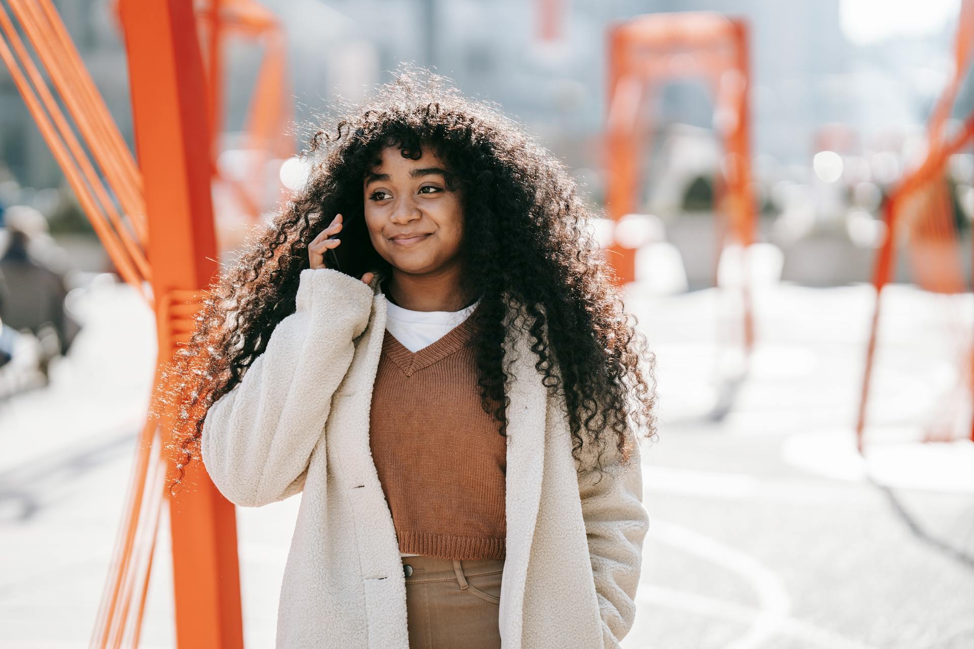 Happy young African American female wearing white coat having pleasant conversation via mobile phone and looking away contentedly while standing on sunny spring city street