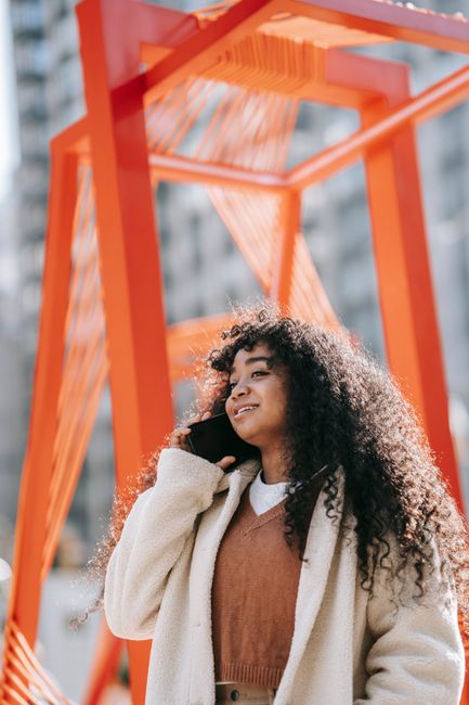 Cheerful young African American female wearing white warm coat chatting on mobile phone while standing on sunny city street and looking away with smile