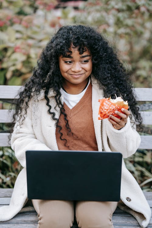 Cheerful African American female wearing warm coat eating yummy burger and browsing netbook on laps while sitting on bench in city park on sunny spring day