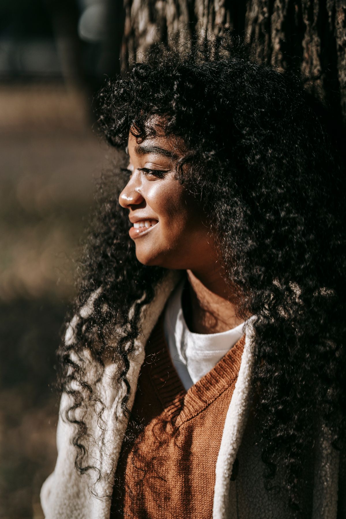 Attractive happy African American female with curly hair leaning on tree in park and looking away during warm spring day