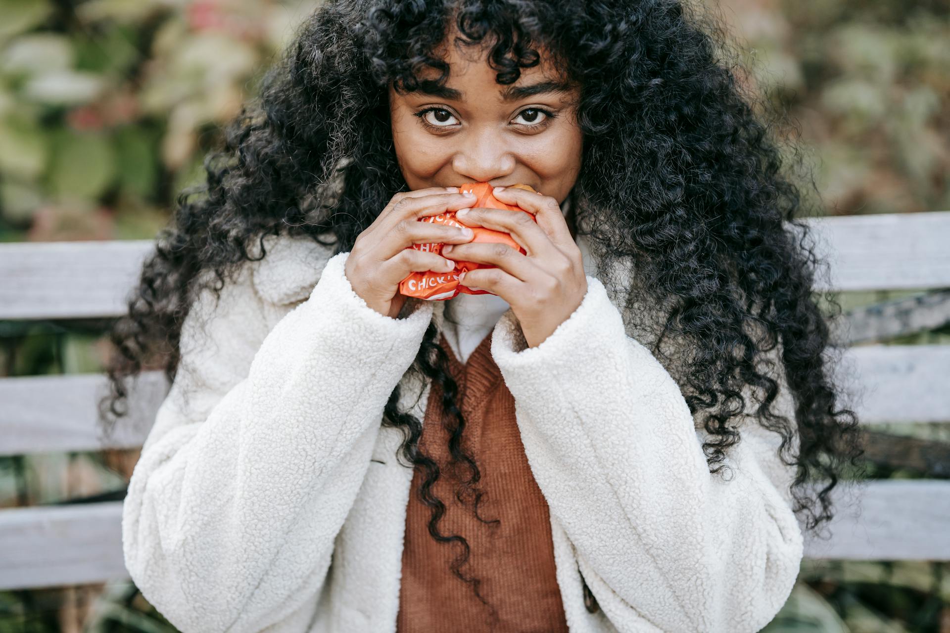 Crop hungry African American female in warm clothes biting delicious fresh burger and looking at camera while sitting on bench in park