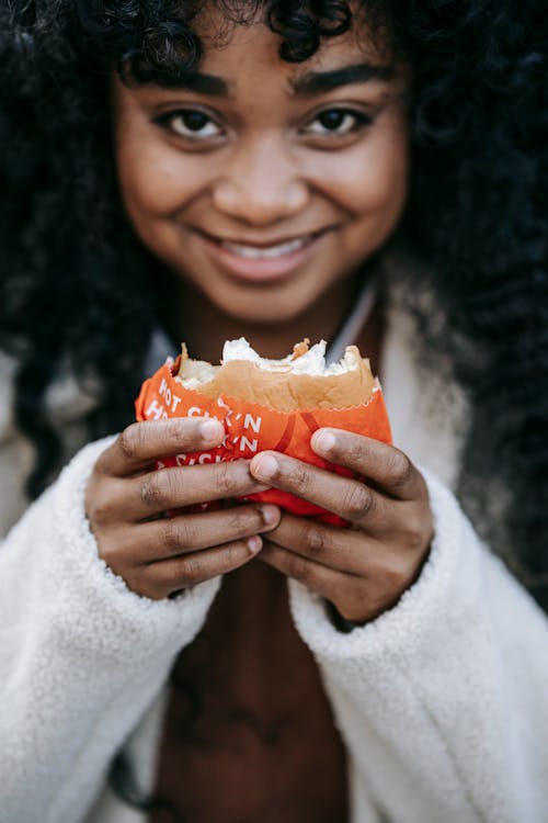 Free Crop smiling African American female wearing white coat eating yummy fresh burger outdoors and looking at camera contentedly Stock Photo