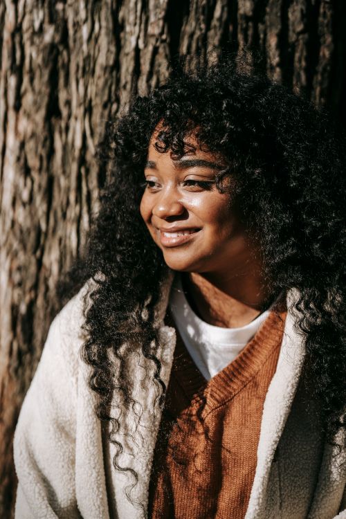 Free Cheerful young African American female wearing casual warm clothes standing against tree in sunny park and looking away with smile Stock Photo