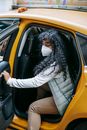 Side view emotionless African American female in warm clothes and face mask opening door and getting off yellow taxi