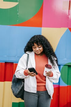 Smiling woman with smartphone and coffee in front of colorful wall.
