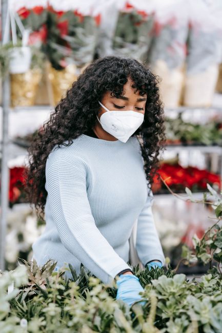 African American female wearing protective mask and gloves picking fresh flowers while standing in floral market with blooming bouquets on blurred background