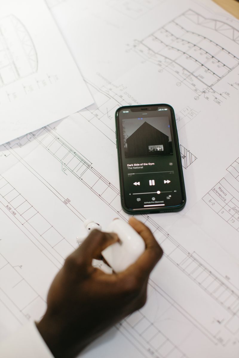 A smartphone playing music on architectural blueprints with a hand holding earbuds.