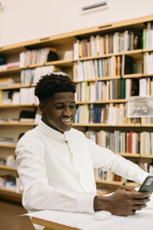 Free Smiling man reading and texting on smartphone in a library setting. Stock Photo