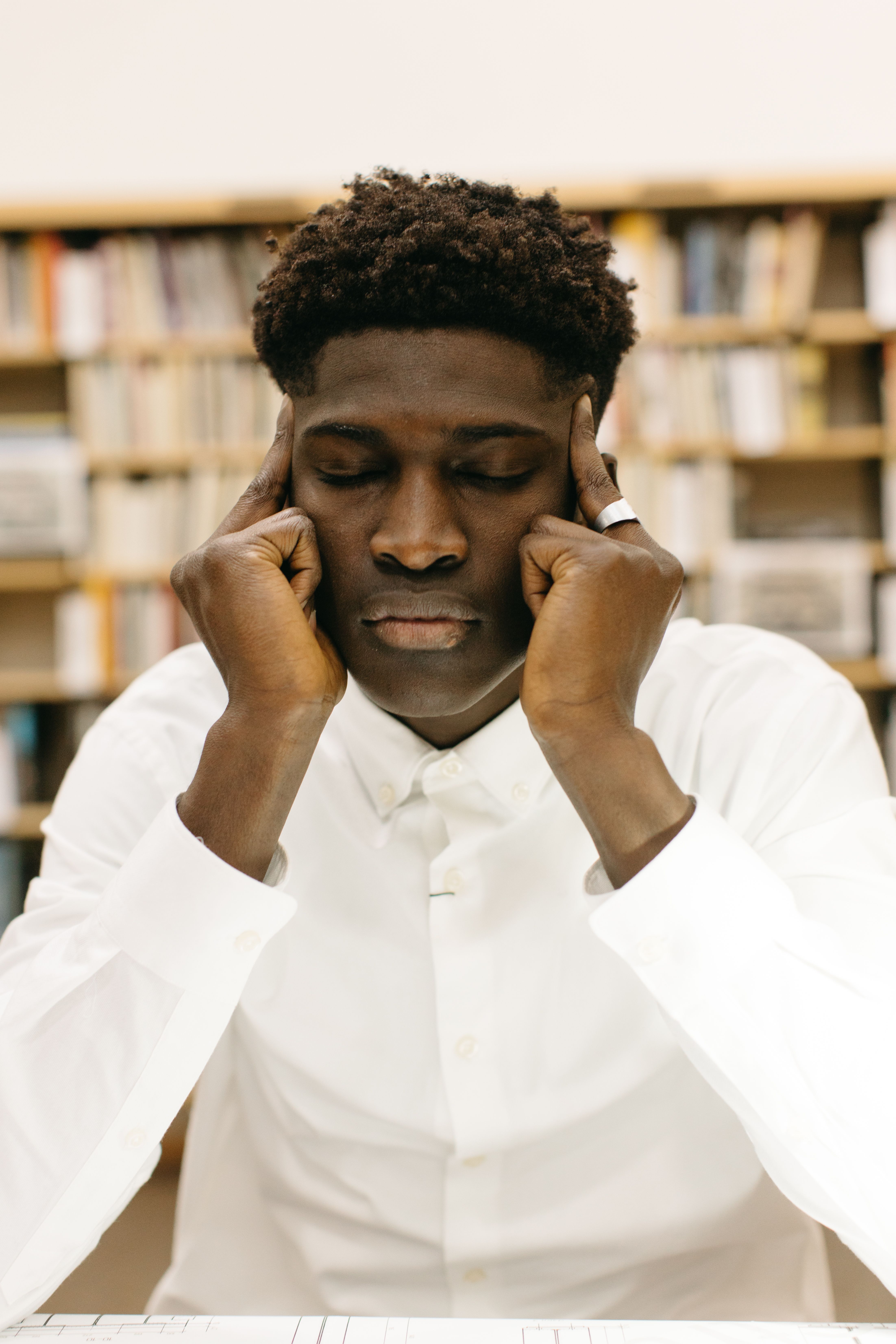 Man in white shirt concentrating in a library, eyes closed, with bookshelves in the background.
