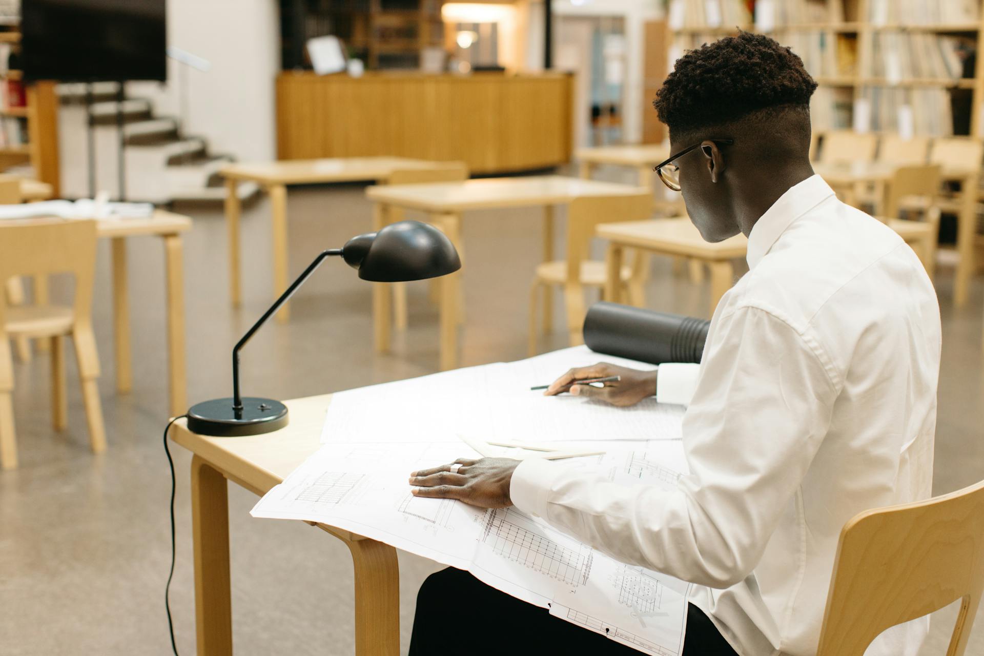 Young man drafting architectural plans at a library table, focused on work.