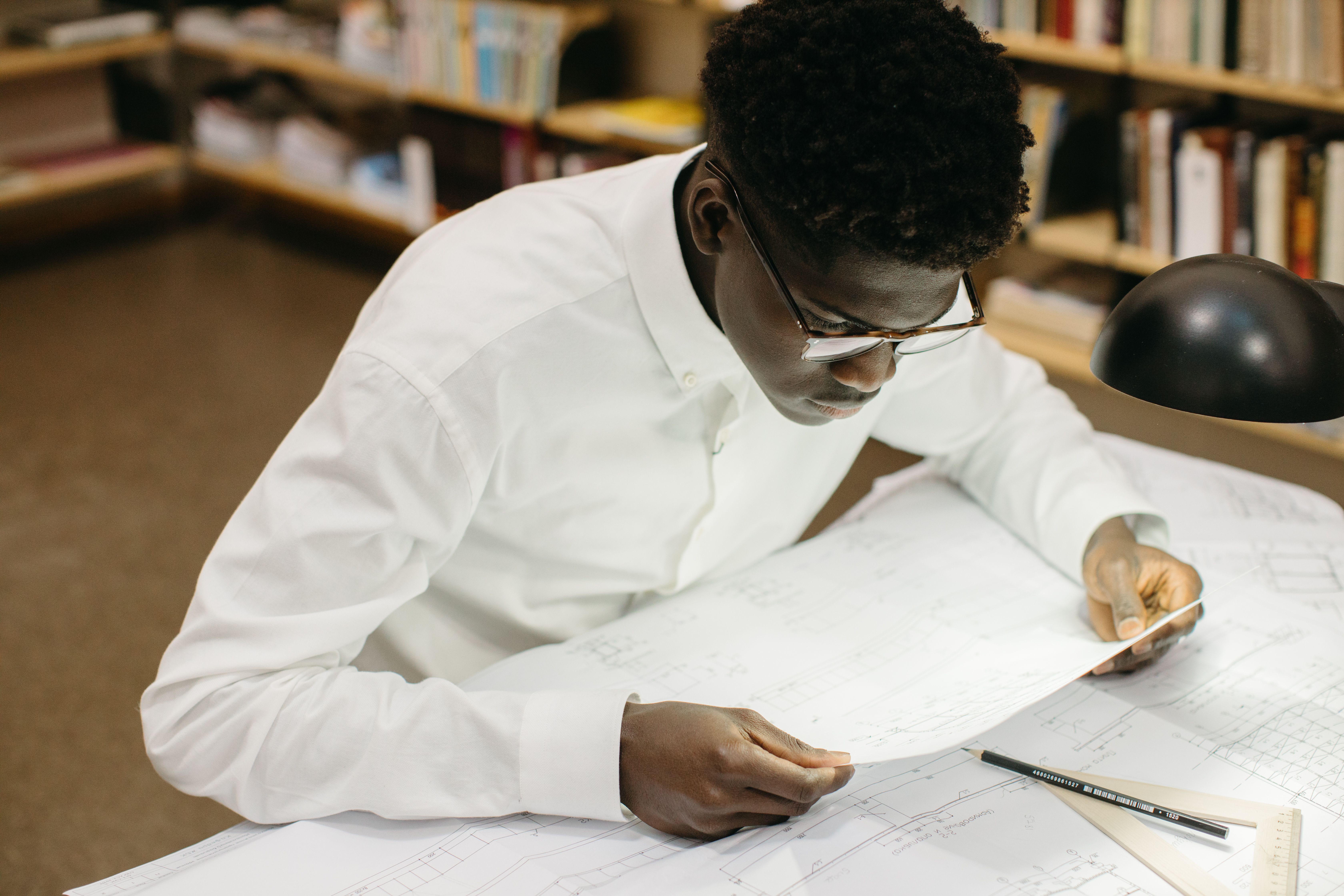 Student wearing glasses reviews architectural plans under a desk lamp in a library