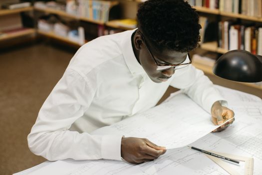 Young man intensely studying architectural blueprints in a library setting.