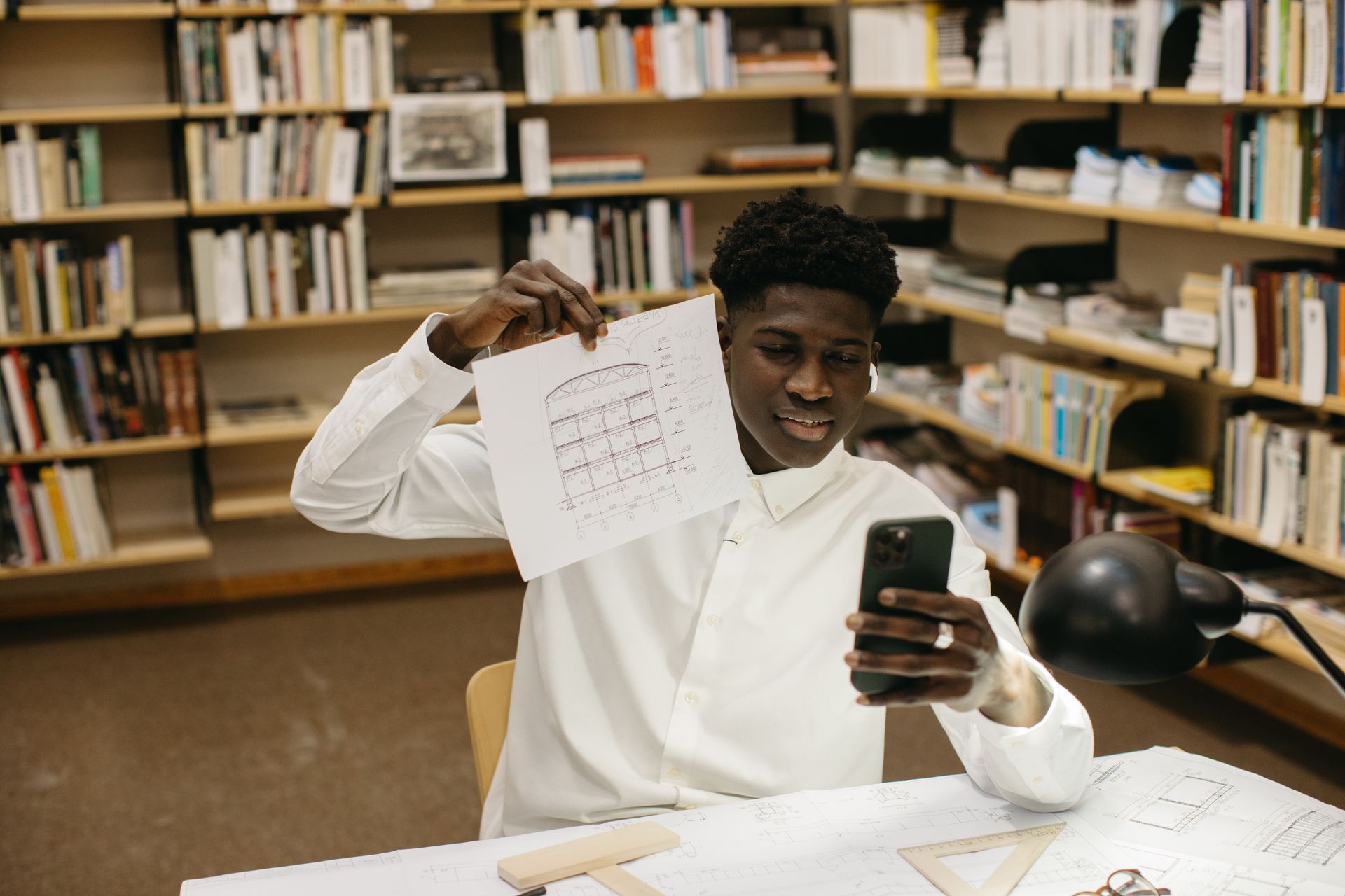 Architect holding a blueprint and phone, smiling in a library workspace.