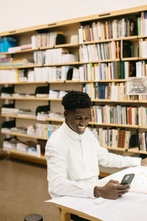 Free Young black man smiling while using smartphone in a library, sitting at a desk with books. Stock Photo