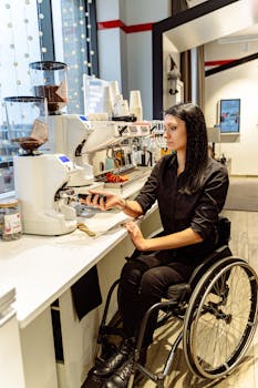 Woman in wheelchair preparing coffee using a grinder in a cafe setting.