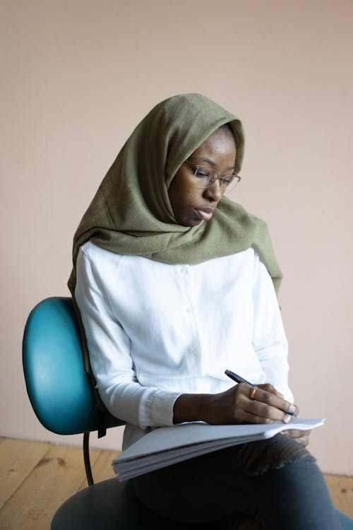 Serious young African American Muslim female wearing casual outfit hijab and eyeglasses taking notes in copybook while sitting on chair in light room