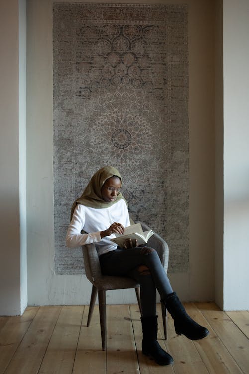 Full body focused African American Muslim female in traditional hijab and casual wear reading interesting story and sitting with legs crossed on armchair in light room