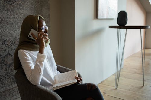 Free Unemotional African American Muslim female in white shirt eyeglasses and hijab sitting on armchair with book and having conversation via mobile phone in modern apartment Stock Photo