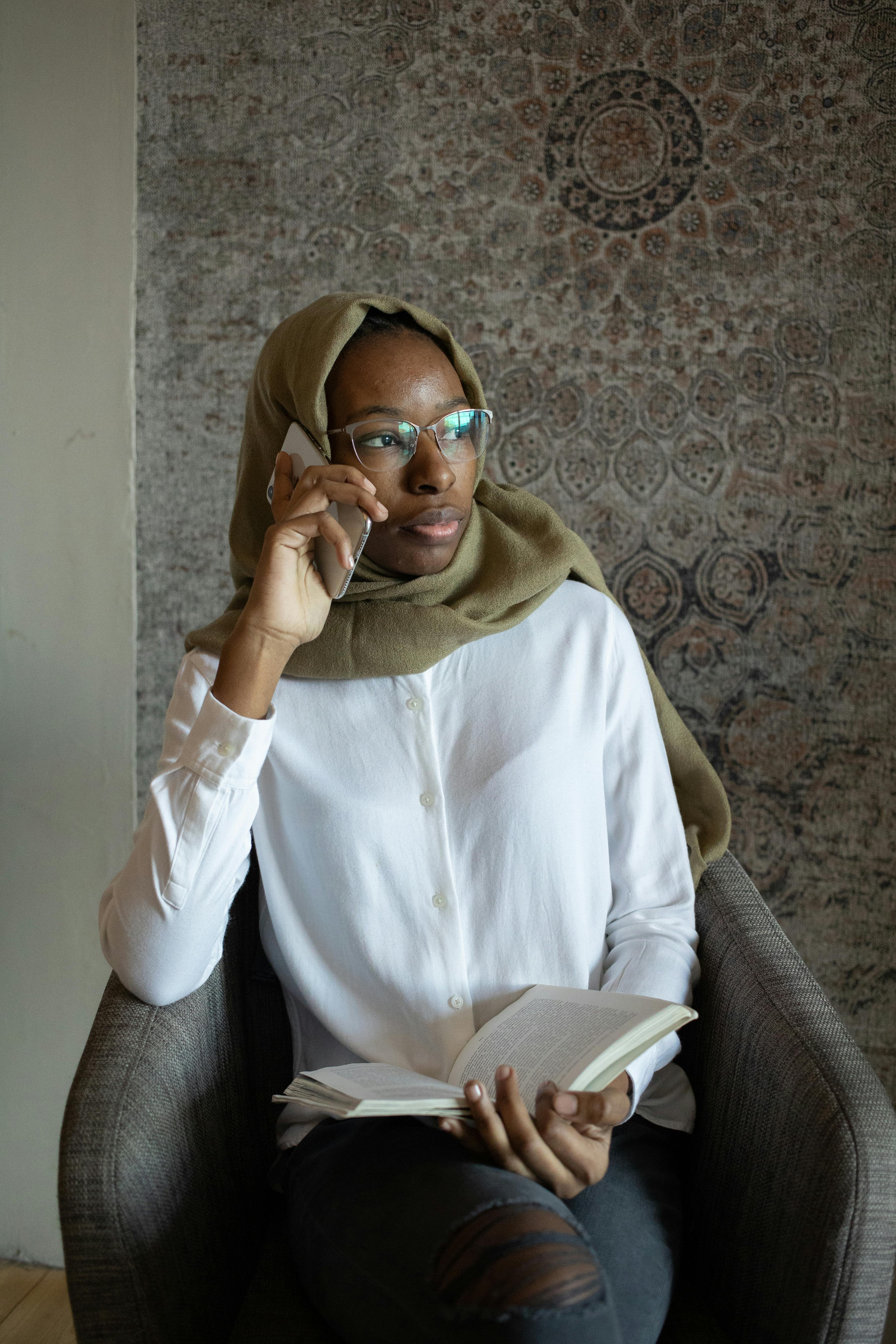 thoughtful black muslim woman with book talking on smartphone