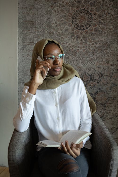 Free Thoughtful black Muslim woman with book talking on smartphone Stock Photo