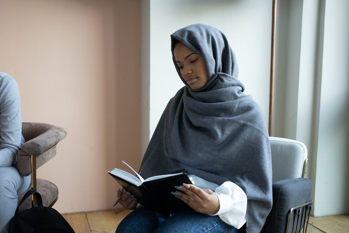 Free Concentrated young Muslim female in traditional headscarf reading novel and sitting on armchair in light room Stock Photo