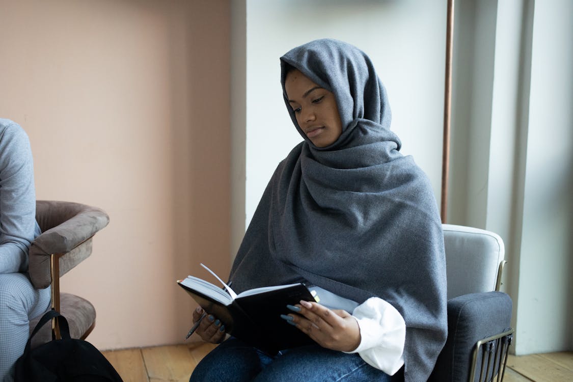 Concentrated young Muslim female in traditional headscarf reading novel and sitting on armchair in light room