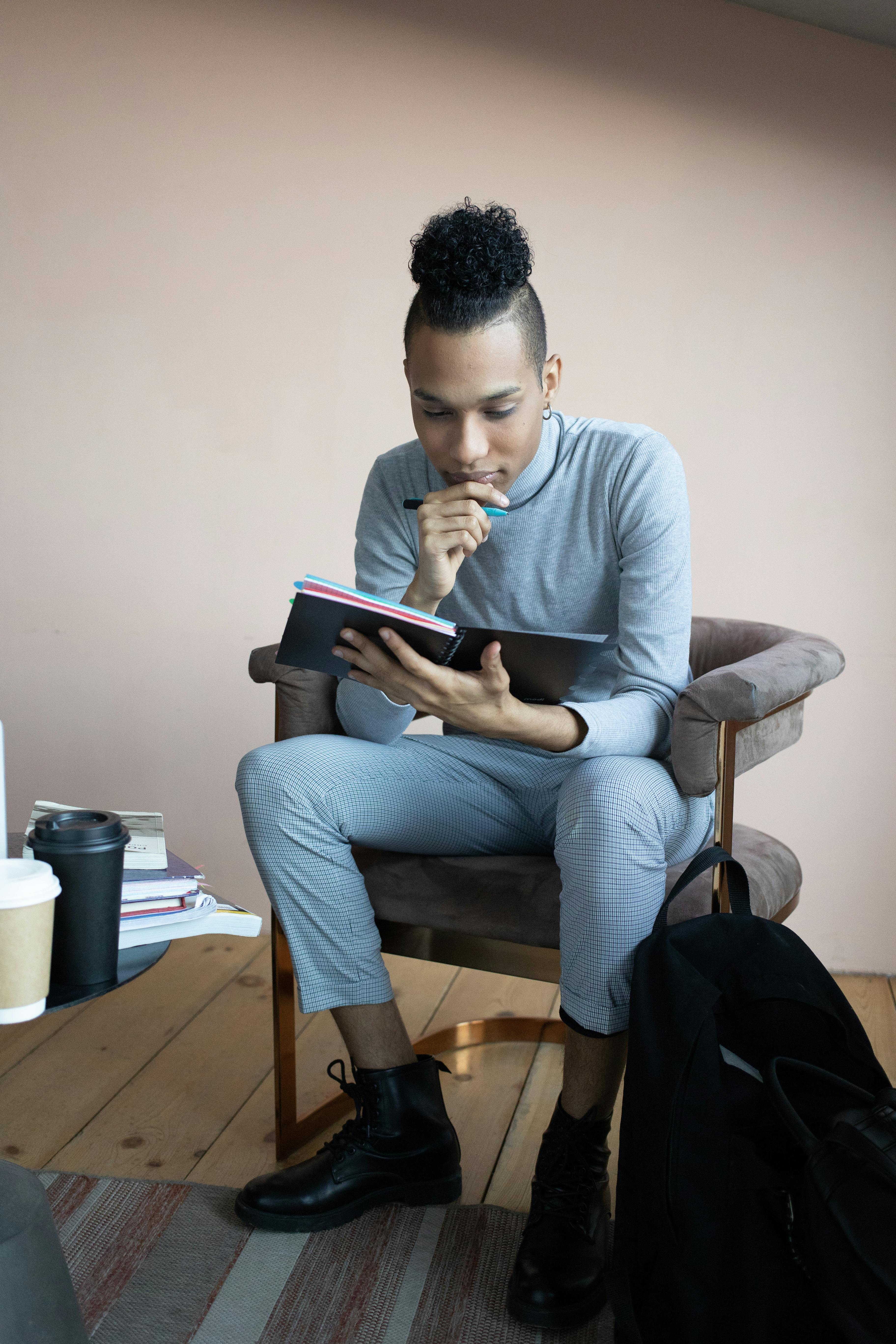 Free A young man sits thoughtfully in a cozy armchair while reading a book indoors. Stock Photo