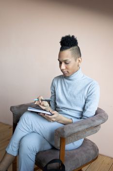 Concentrated young ethnic male in casual outfit sitting with pen and notebook on comfortable armchair in light studio