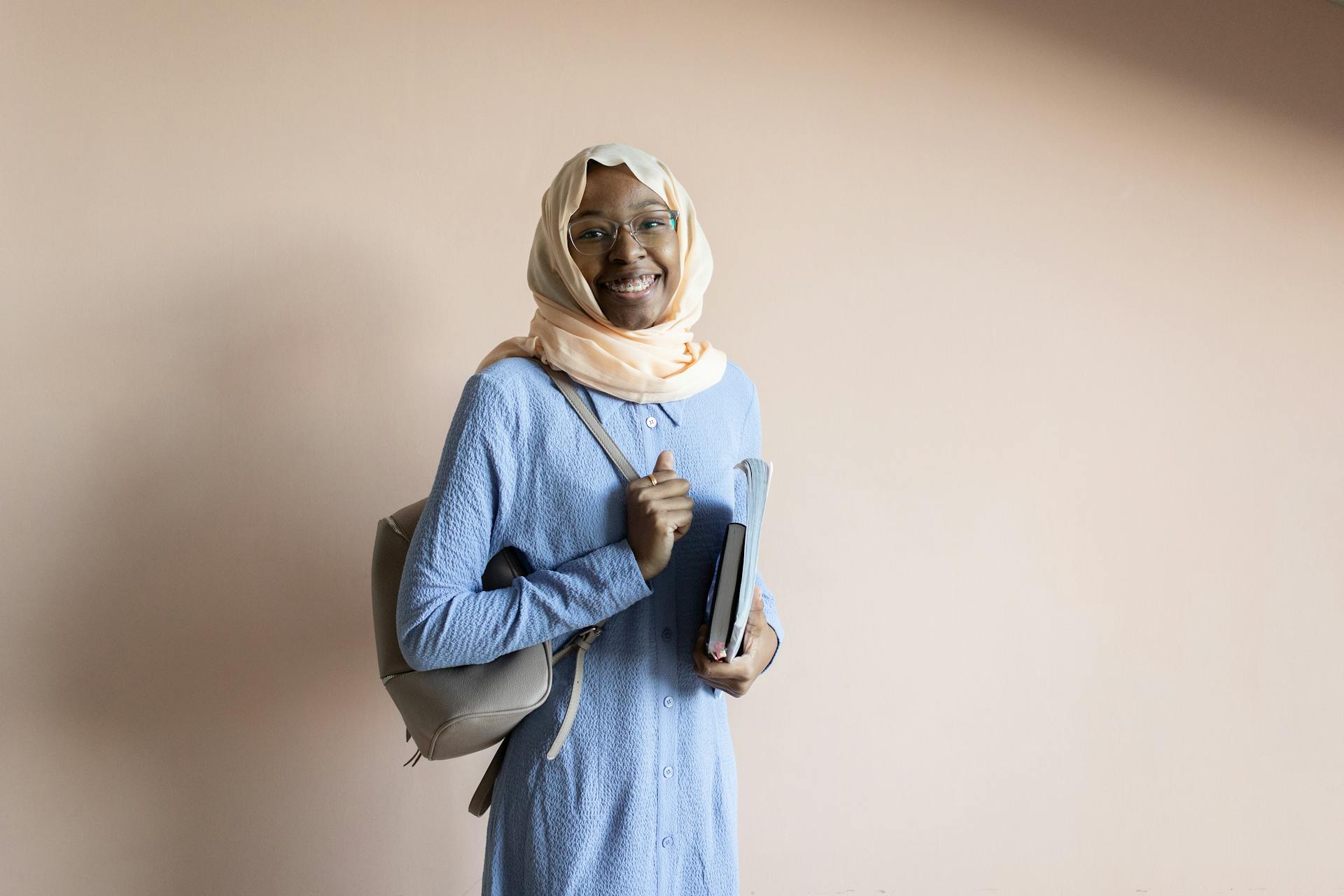 Smiling Muslim woman in hijab holding a book and backpack, symbolizing education and confidence.