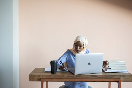 A Muslim woman in a headscarf studies diligently using a laptop at her desk with books and a cup.