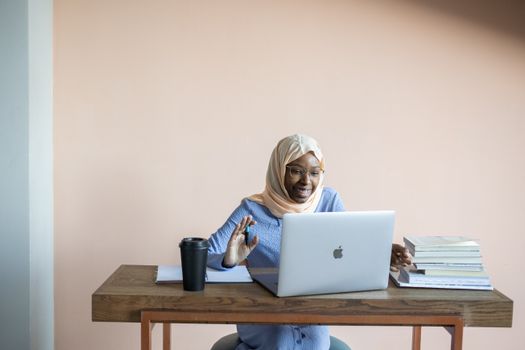 Positive African American female in headscarf sitting at table with books and cup and waving hand to laptop during remote lesson