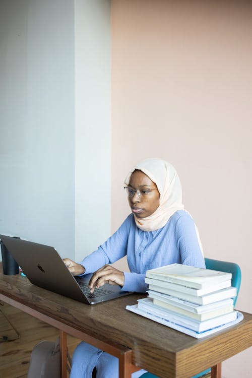Free Concentrated African American female student in hijab studying on netbook sitting at table with stack of books Stock Photo