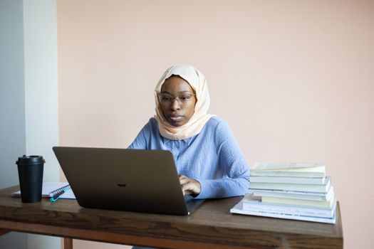 Concentrated African American female in hijab and eyeglasses browsing netbook sitting at table with stack of books and reusable cup