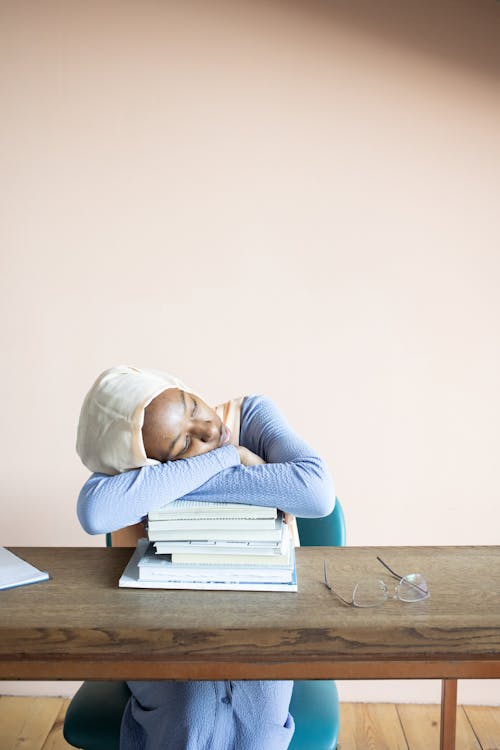 Free Exhausted African American female student in hijab having nap on stack of textbooks after studies Stock Photo